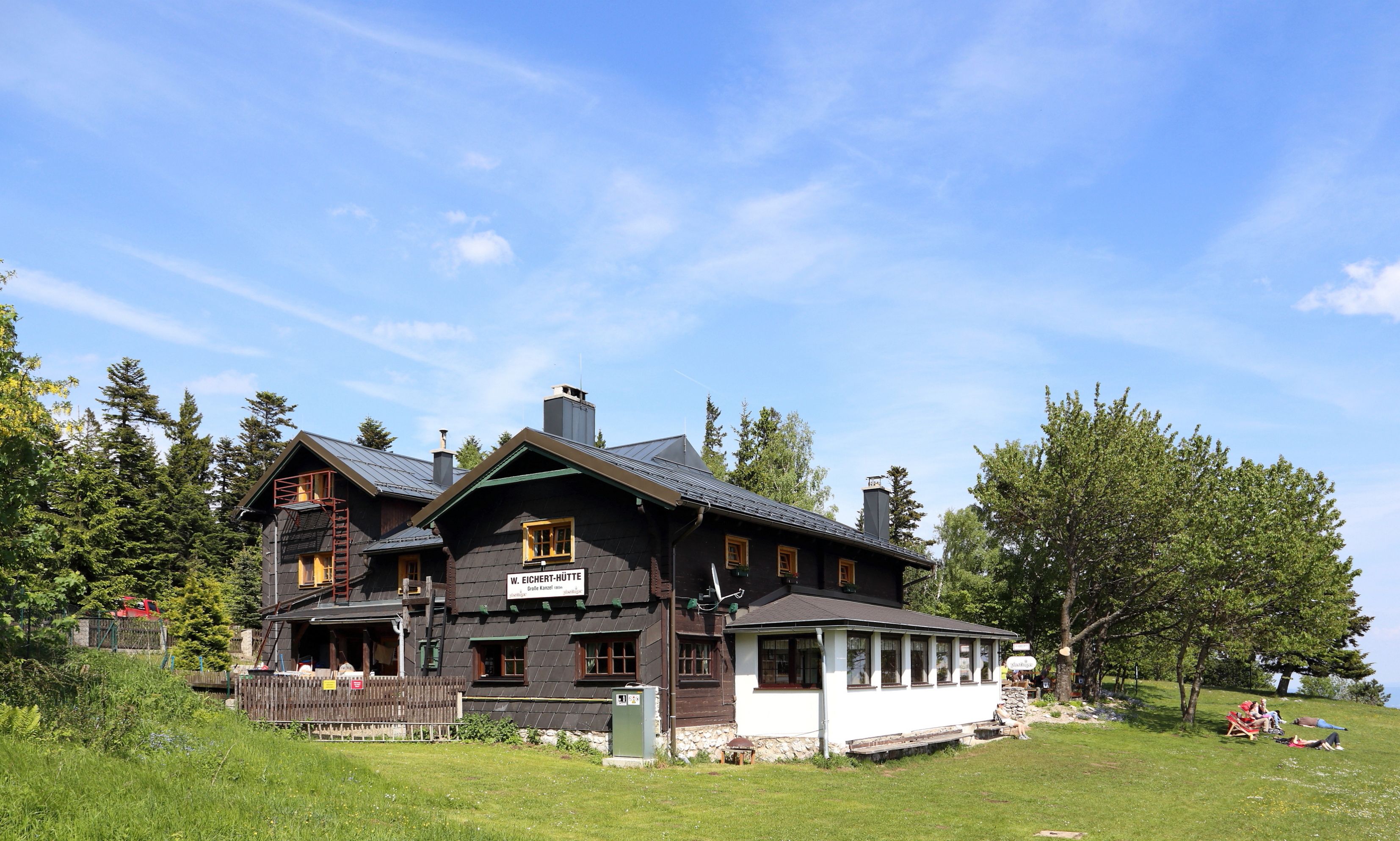 A mountain hut with dark wood and white windows, surrounded by trees and meadows, under a blue sky.