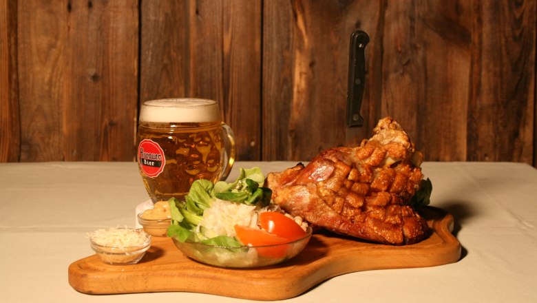 A wooden board with pork knuckle, salad and beer mug in front of a wooden wall.