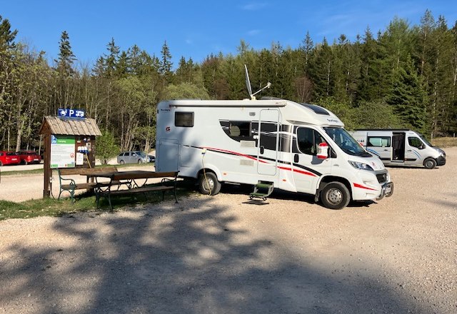Wohnmobil auf einem Parkplatz im Naturpark Hohe Wand, umgeben von B&auml;umen und einem blauen Himmel.
