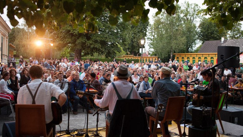 Open-air concert with musicians on a stage and a large outdoor audience.